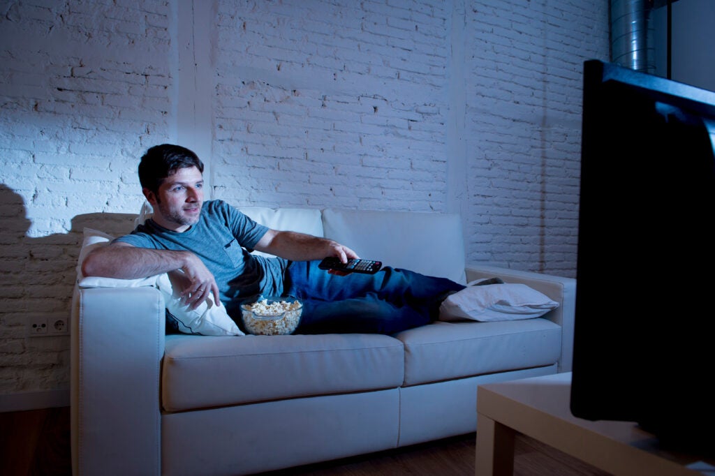 man at home lying on couch in living room with popcorn watching tv holding remote control.