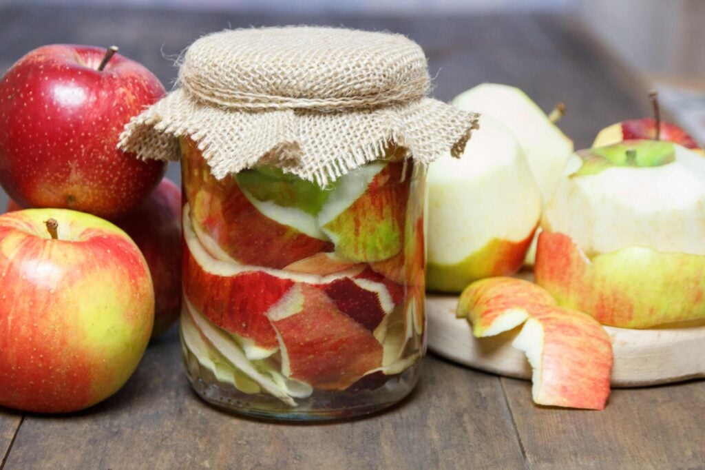 Apples (some unpeeled, some peeled) next to a small fabric-covered jar of apple peels in liquid.