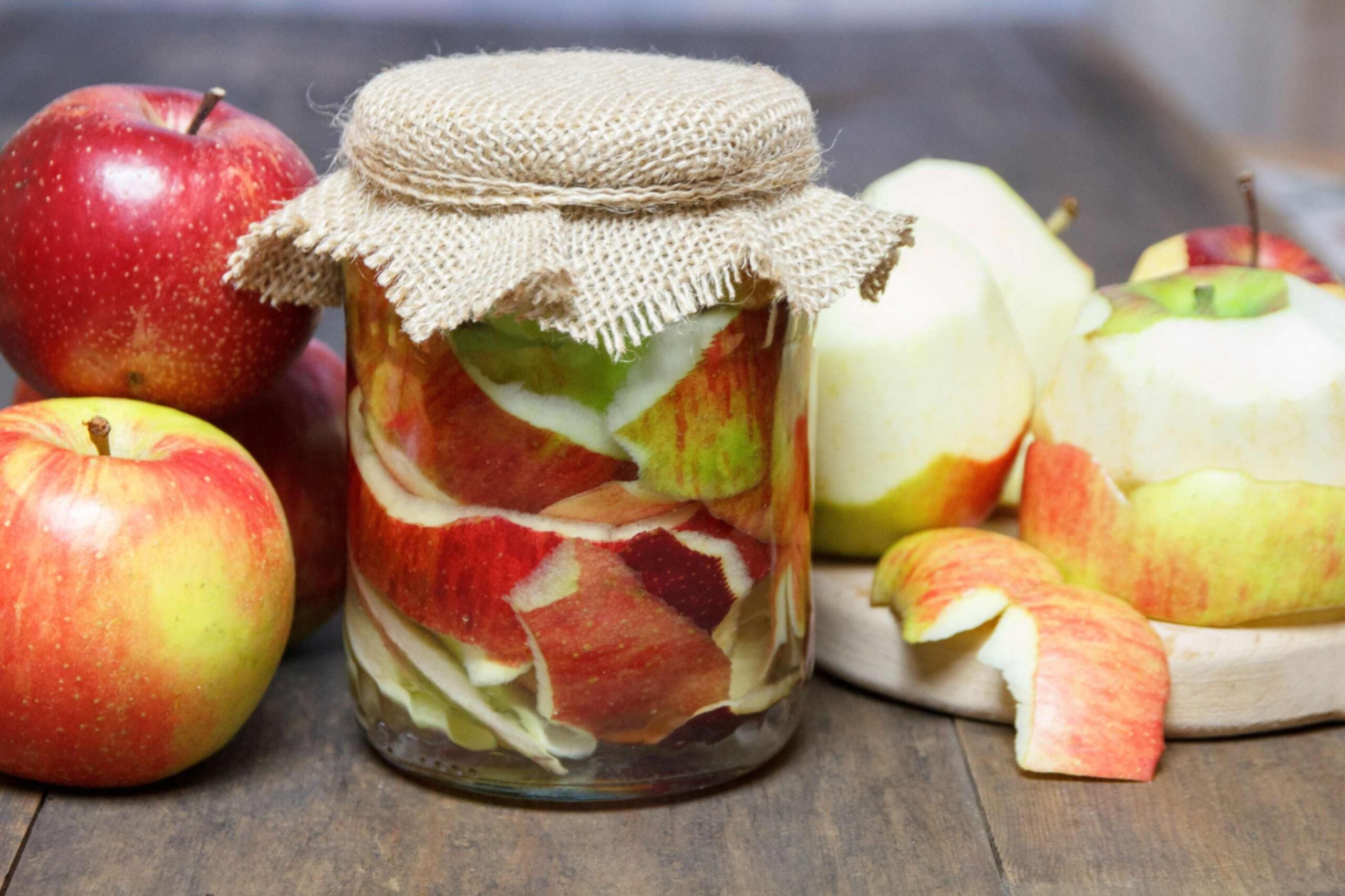 Apples (some unpeeled, some peeled) next to a small fabric-covered jar of apple peels in liquid.
