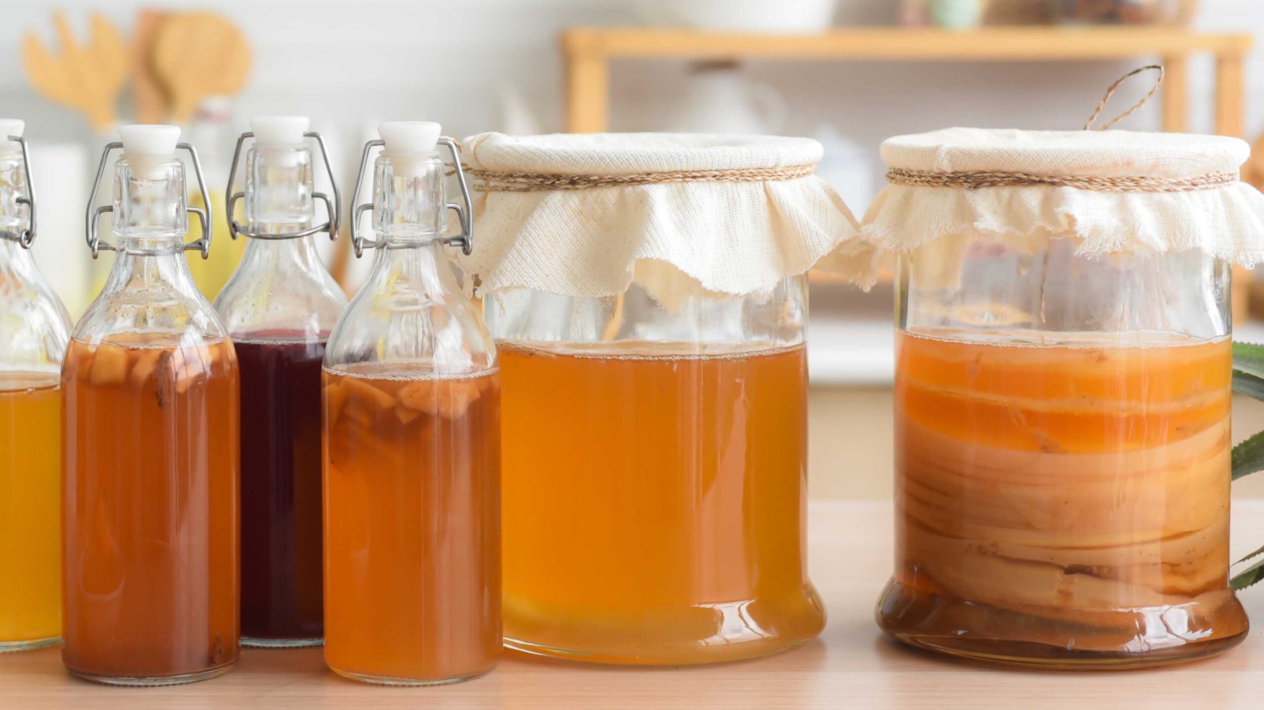 glass bottles of different colors of Kombucha, next to two bigger jars with fabric over the tops