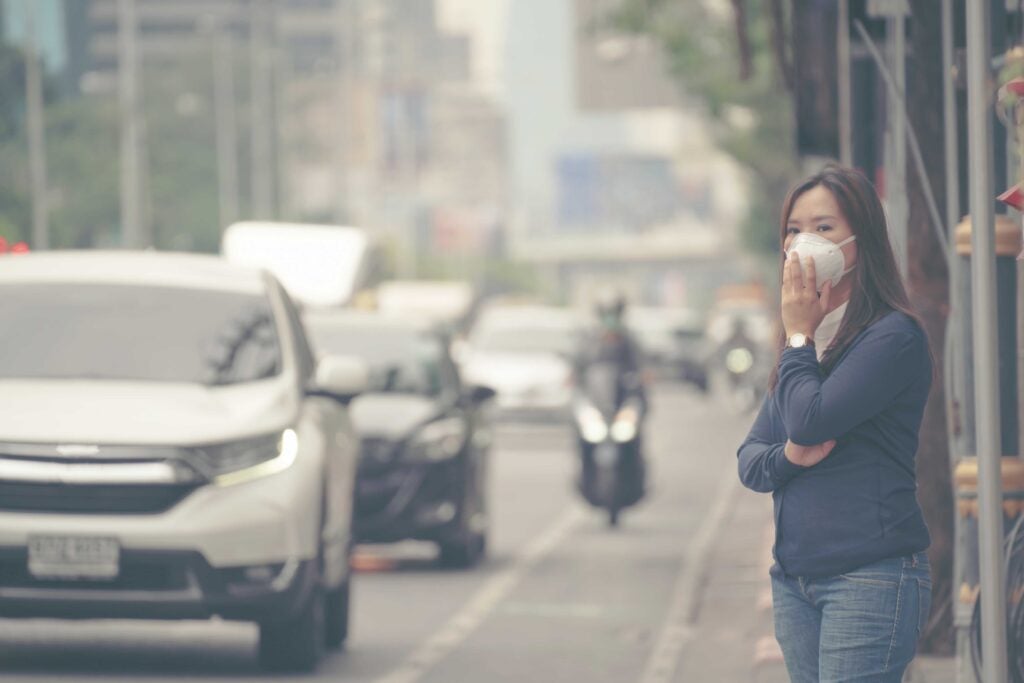 Masked woman standing on side of road in a smoggy area.